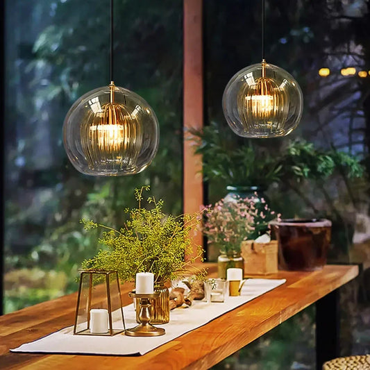 Un par de lámparas colgantes de vidrio en forma de esfera moderna cuelgan sobre la mesa del comedor, con una luz suave, mientras el fondo muestra un paisaje exterior lleno de verde. Sobre la mesa hay flores y velas, creando un ambiente acogedor.